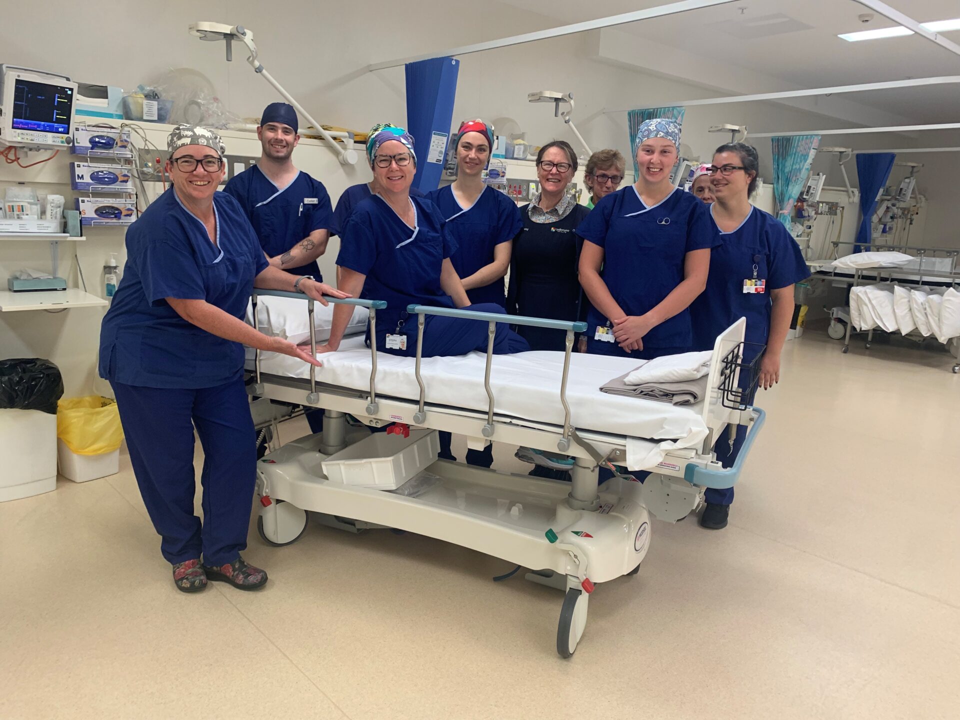 A group of eight theatre nurses stand around the new trolley. They are smiling. The photo is taken in the Operating Theatre recovery room.