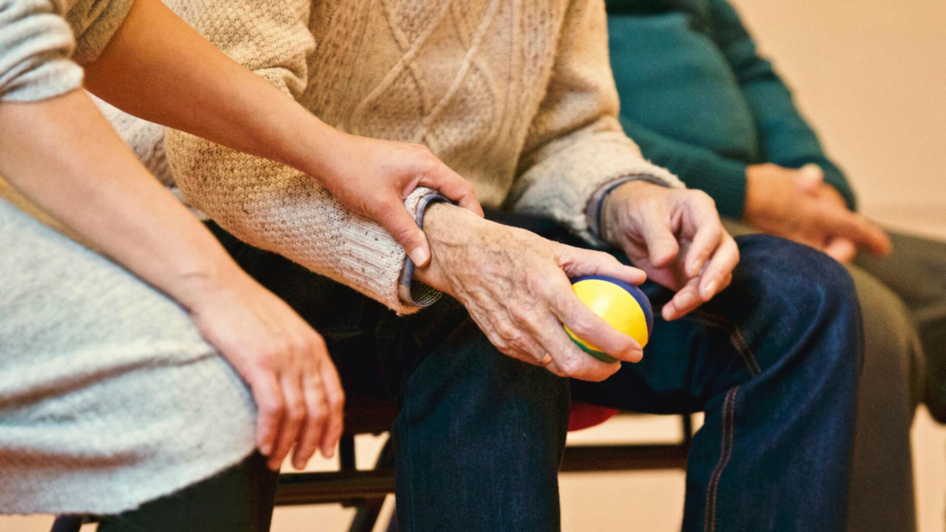 Three people sitting in a group in a circle, only their hands and arms are visible. One hand is holding an older man's arm in a comforting way