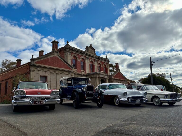 a range of classic cars are parked in front of the historic facade of the Maldon Hospital with the background of a few clouds in a blue sky.