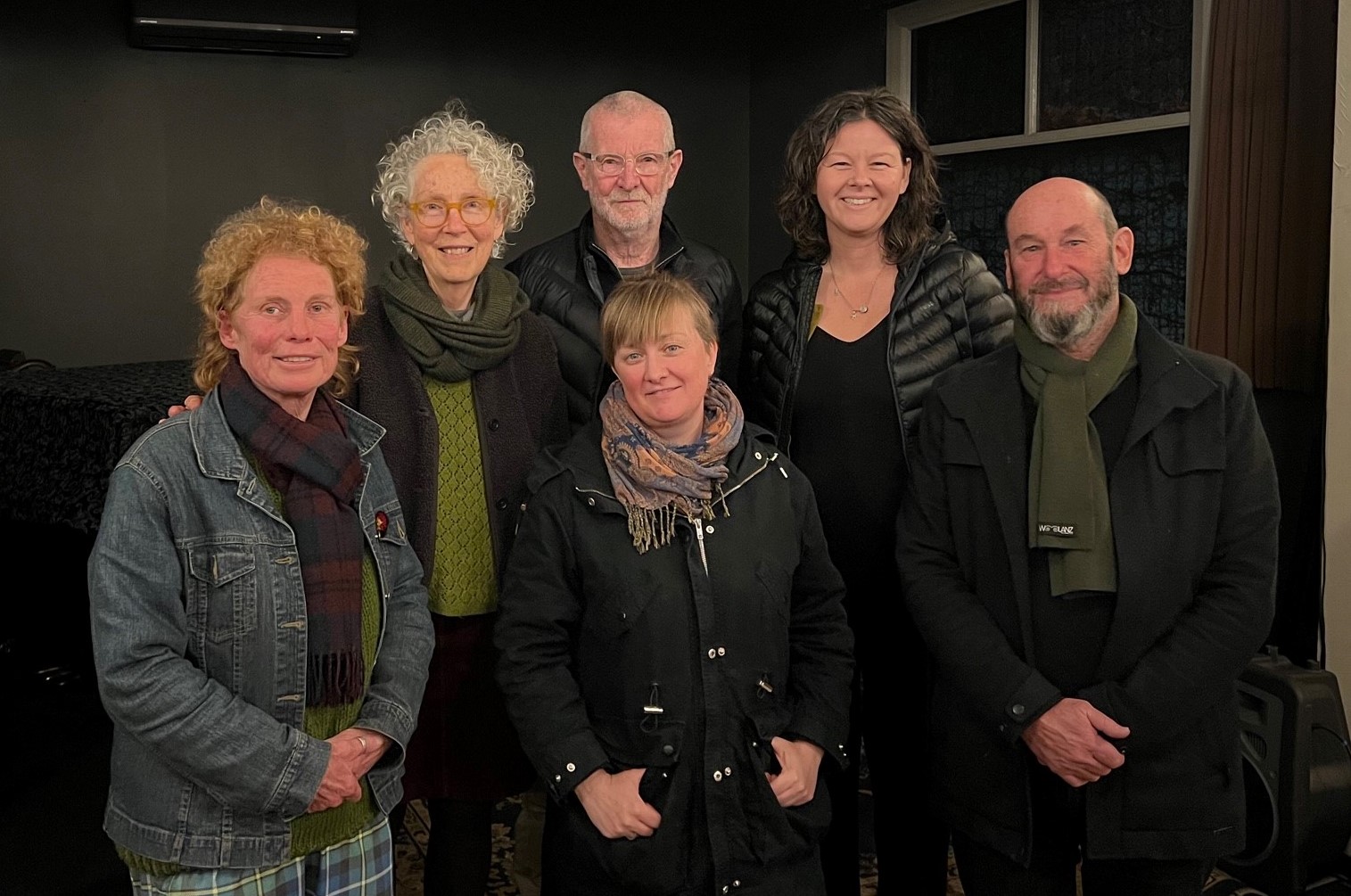 4 women and two men, all smiling and dressed in warm dark clothing in a room with low light
