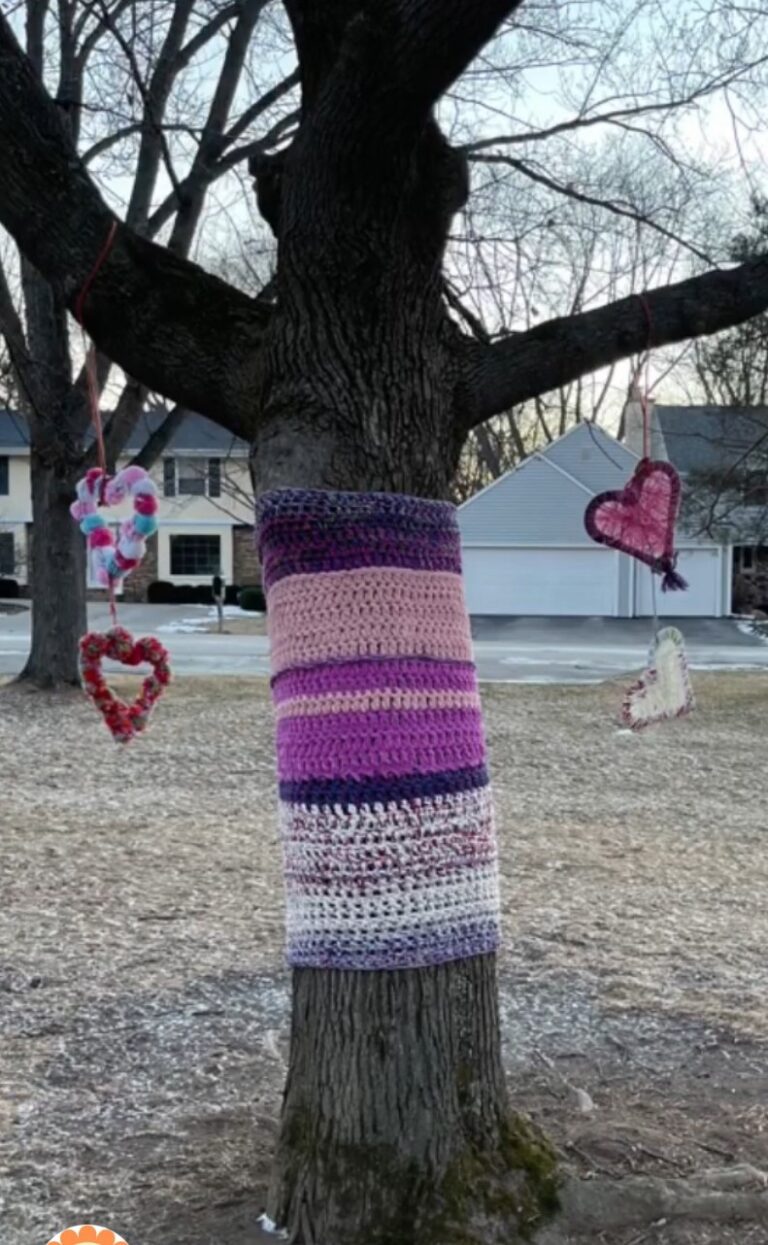 a bare tree with it's trunk wrapped in purple and pink striped knitting. There are woollen crafted hearts hanging from the lower branches