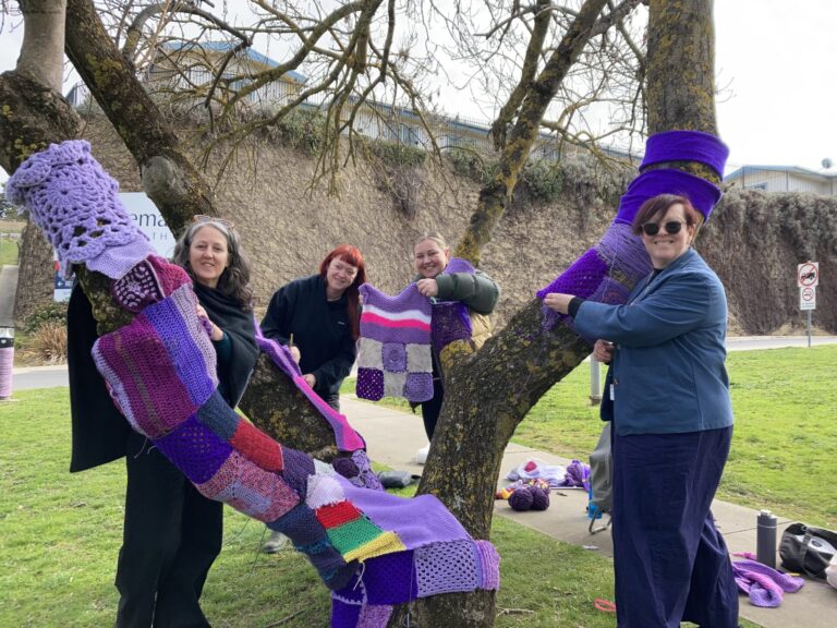 Gen, Bethia, Robyn & Sherene attaching knitted and crocheted purple pieces to a bare-leafed tree