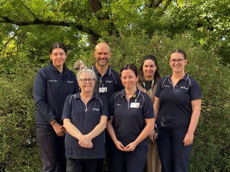 six smiling staff in Dhelkaya Health navy blue uniforms standing in front of a green leafy tree