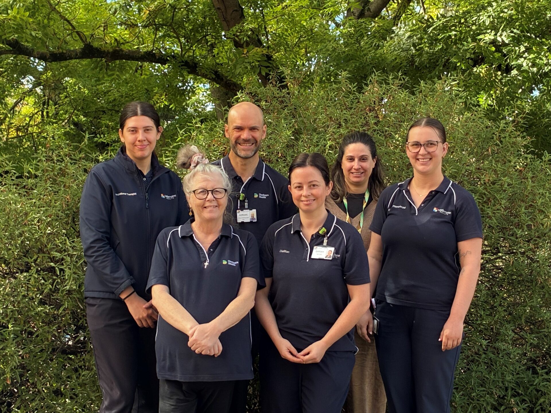 six smiling staff in Dhelkaya Health navy blue uniforms standing in front of a green leafy tree