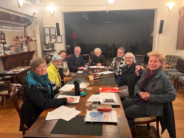 8 adults meeting around a long wooden table, all dressed in warm casual clothes