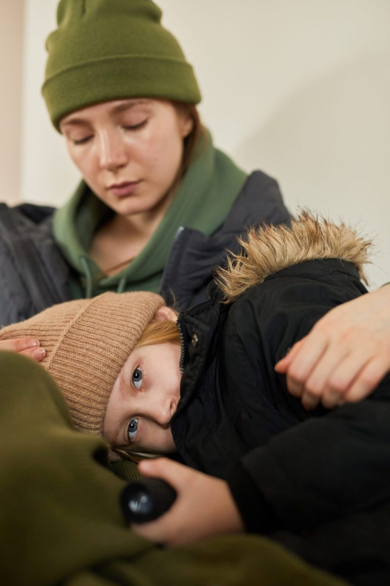 a mother sitting on the ground with her young daughter laying with head in mum's lap, both rugged up against the cold