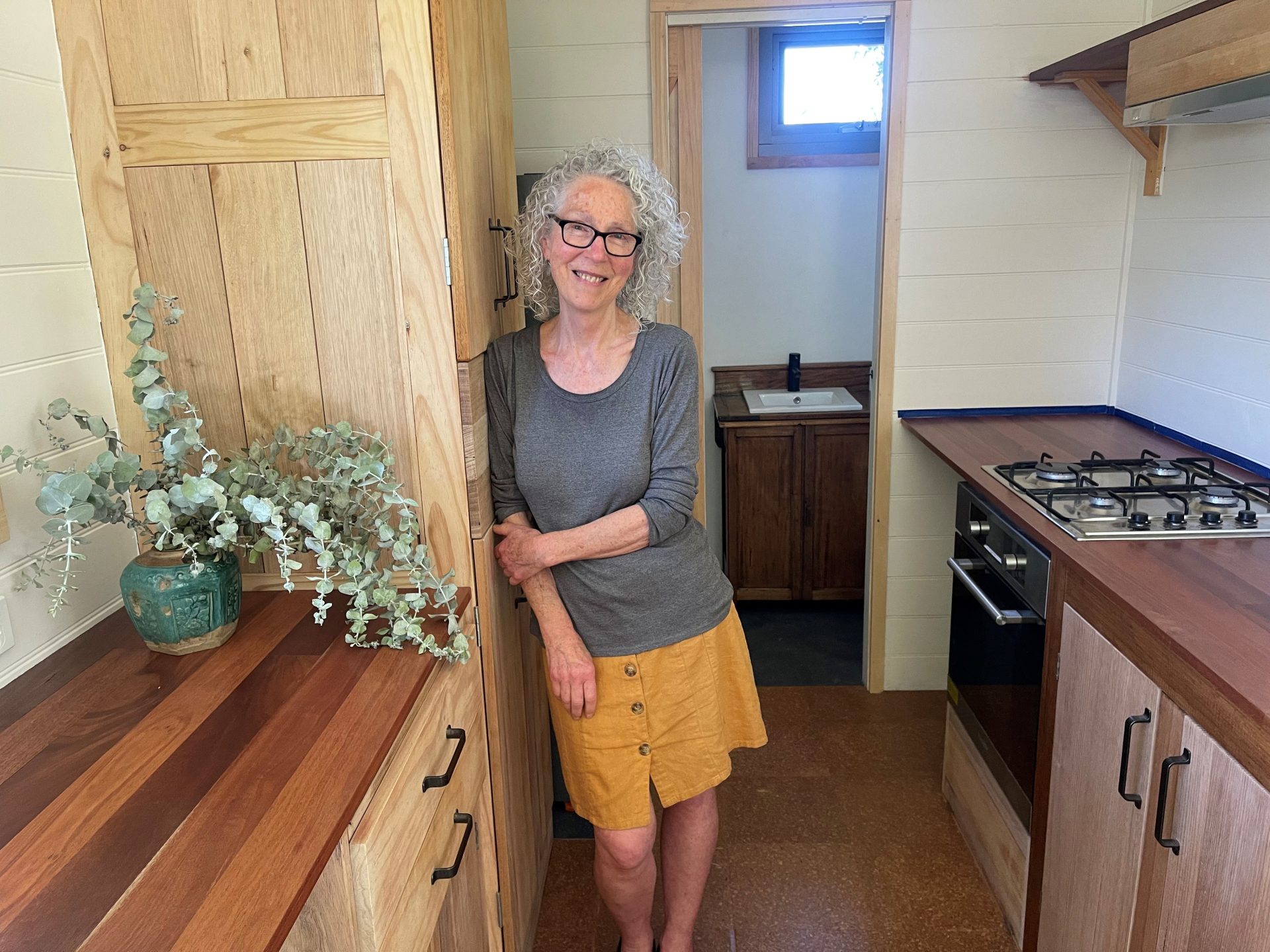 a woman leaning against a cupboard inside the kitchen area of her tiny home on wheels.